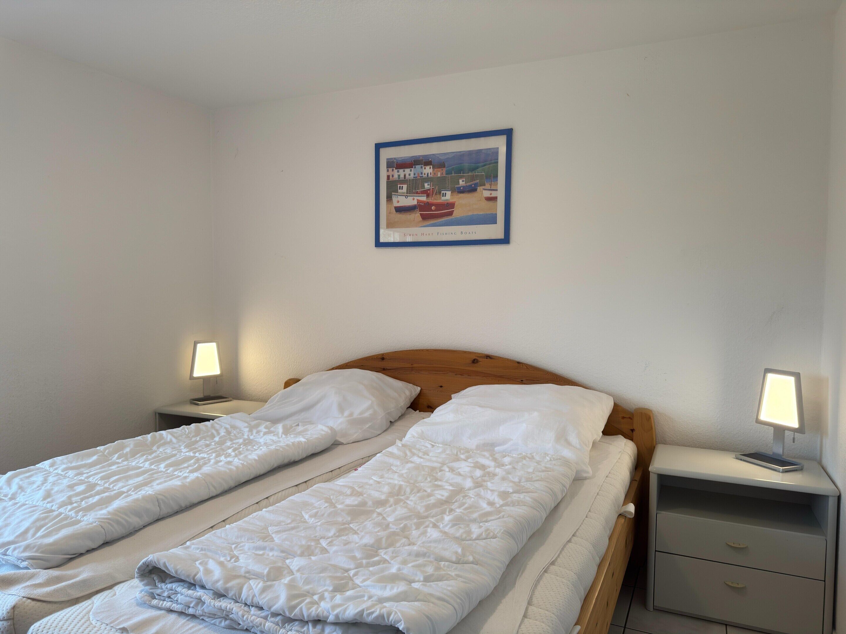 A double bed with white bedding, a wooden headboard, two bedside tables with lamps, and a framed picture of boats on the wall.