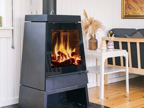 Un estufa de leña negra con puerta de cristal, una mesa blanca con un florero y velas, y un banco de madera en una habitación con paredes blancas y suelo de madera.