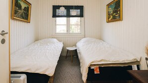 A room with two single beds, a small table, a window with blinds, and two framed pictures on the walls.