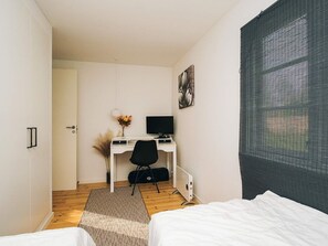 Un dormitorio con cama, escritorio con computadora, silla y ventana con mosquitero.