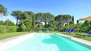 Une piscine d’un bleu éclatant, bordée d’une terrasse carrelée et entourée de haies et d’arbres verdoyants.