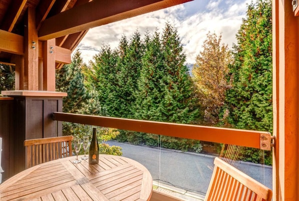 Eine Holzterrasse mit rundem Tisch und Stühlen, mit Blick auf eine Straße und dichtes Grün.