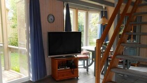 A room with a television on a wooden stand, a clock on the wall, and a staircase. - Ferienhaus im Ferienpark Mirow (Mirow)