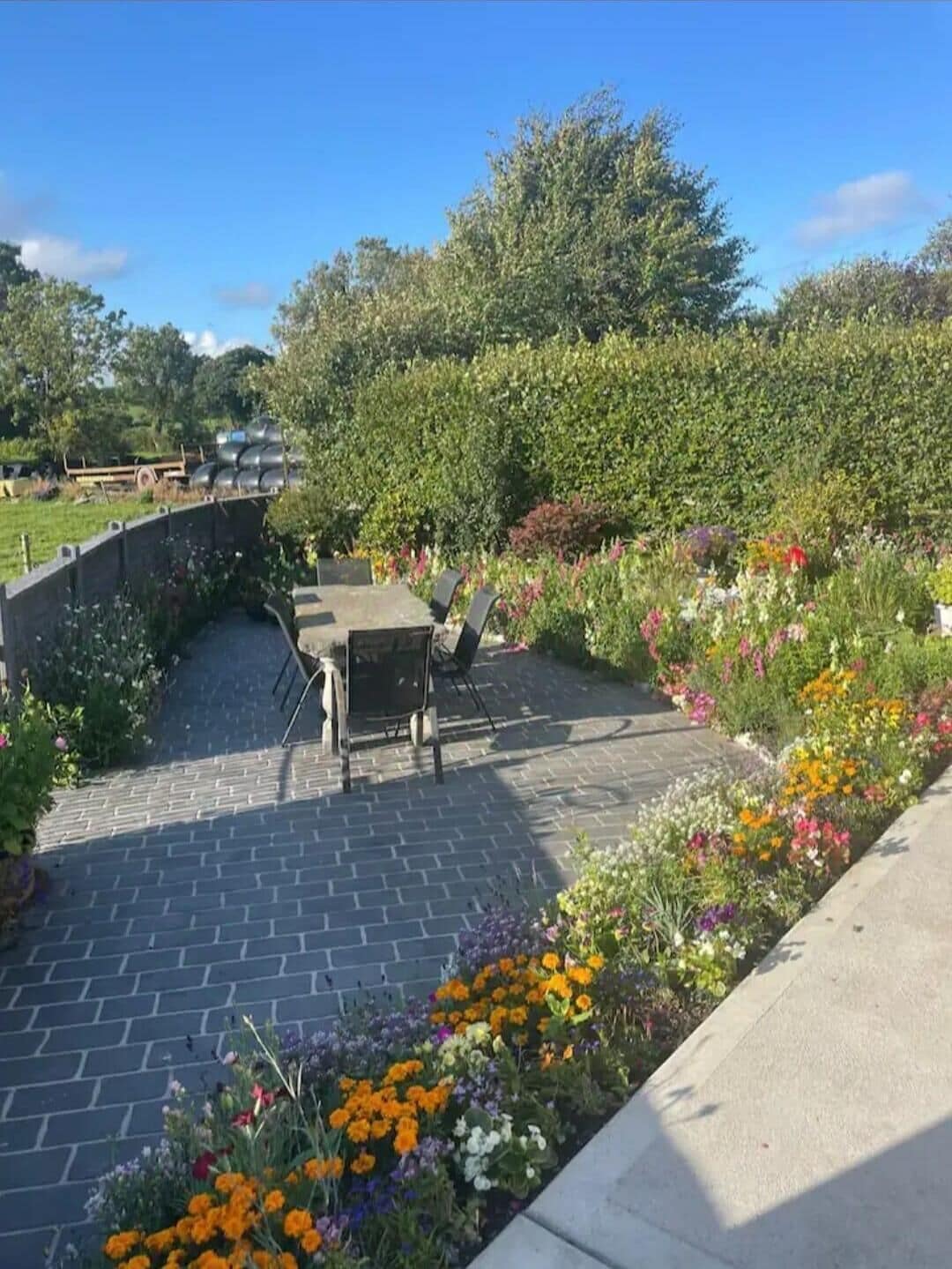 Une terrasse dallée, agrémentée d’une table et de chaises, et entourée d’une variété de fleurs et de plantes aux couleurs vives.