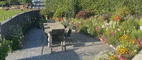 A paved patio area with a table and chairs, surrounded by a variety of colorful flowers and plants.