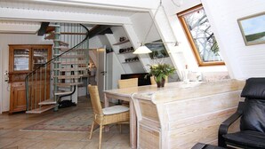 A cozy living room with a wooden dining table, a black leather sofa, and a wicker chair. A staircase with a wooden railing leads to an upper level.