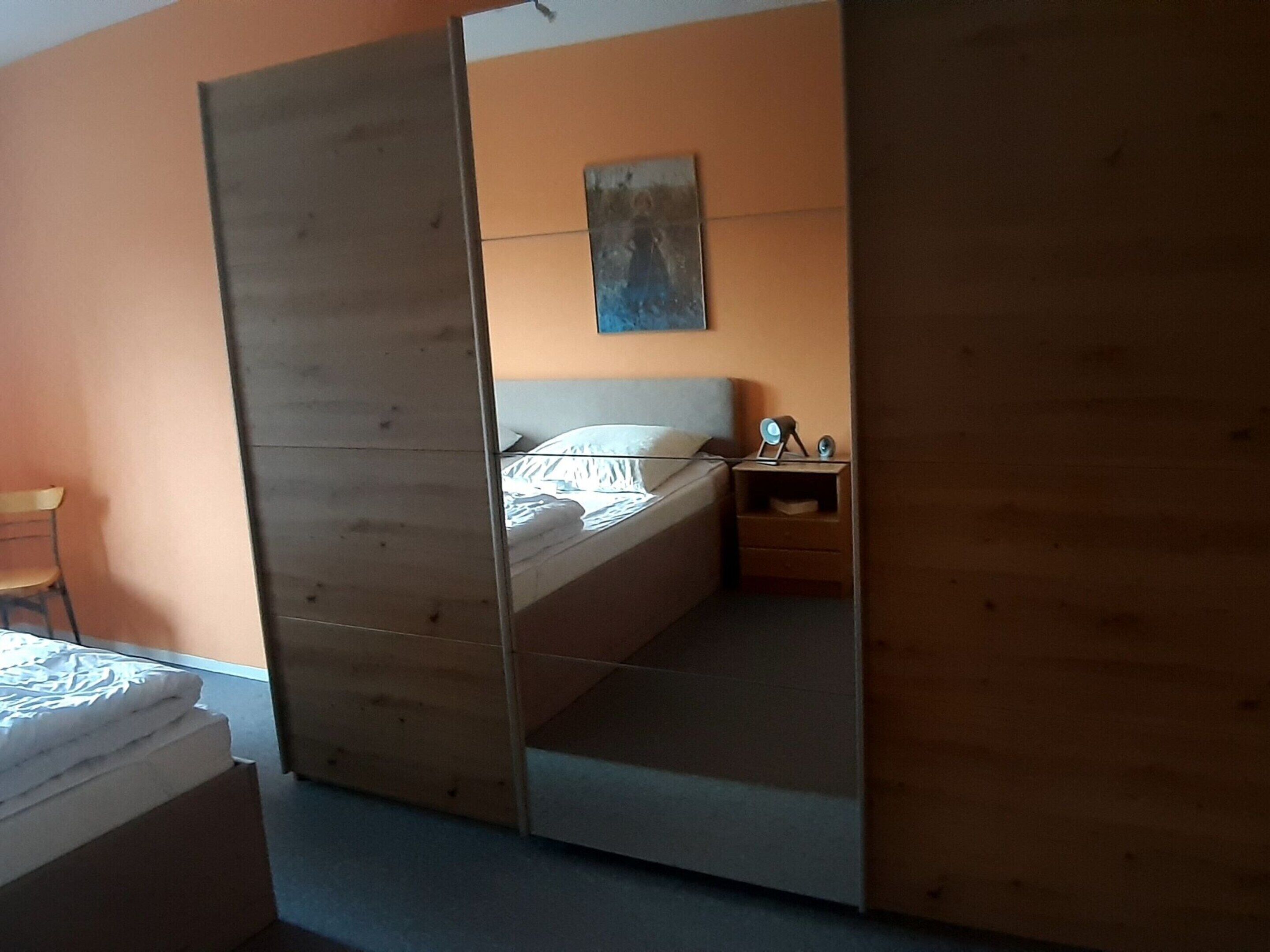A bedroom with a wooden wardrobe, a bed with white bedding, a nightstand with a lamp, and a painting on the wall.