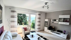 Ein modernes Wohnzimmer mit einem weißen Sofa, einem Couchtisch und einem Fernseher auf einem weißen Schrank. Ein großes Fenster mit Vorhängen, Blick ins Grüne und ein Deckenventilator.