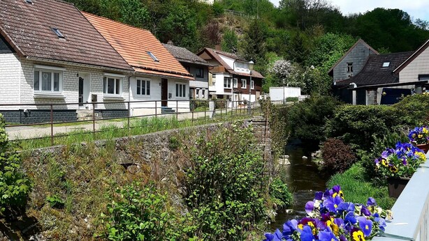 Eine Reihe traditioneller Häuser mit geneigten Dächern, eine Steinmauer und ein kleiner Bach mit Blumen.