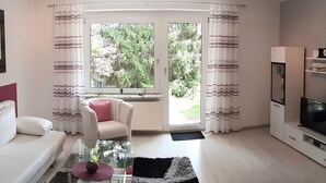 Ein modernes Wohnzimmer mit Sofa, Sesseln, einem Glastisch und einem Fernseher auf einem Schrank.