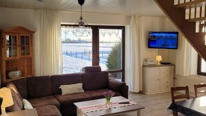 Una sala de estar con un sofá marrón, una mesa de centro blanca, un televisor en la pared y vistas a un paisaje nevado a través de amplios ventanales.