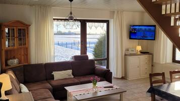 A living room with a brown sofa, a white coffee table, a TV on the wall, and a view of a snowy landscape through large windows.