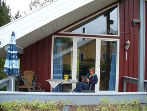 Ein überdachter Außenbereich mit Tisch und Stühlen, eine Person liest ein Buch, und eine Glastür führt nach innen.