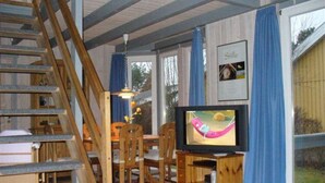 Ein Zimmer mit einer Holztreppe, einem Fernseher auf einem Holzständer, einem Esstisch mit Stühlen und einem mit Vorhängen ausgestatteten Fenster.