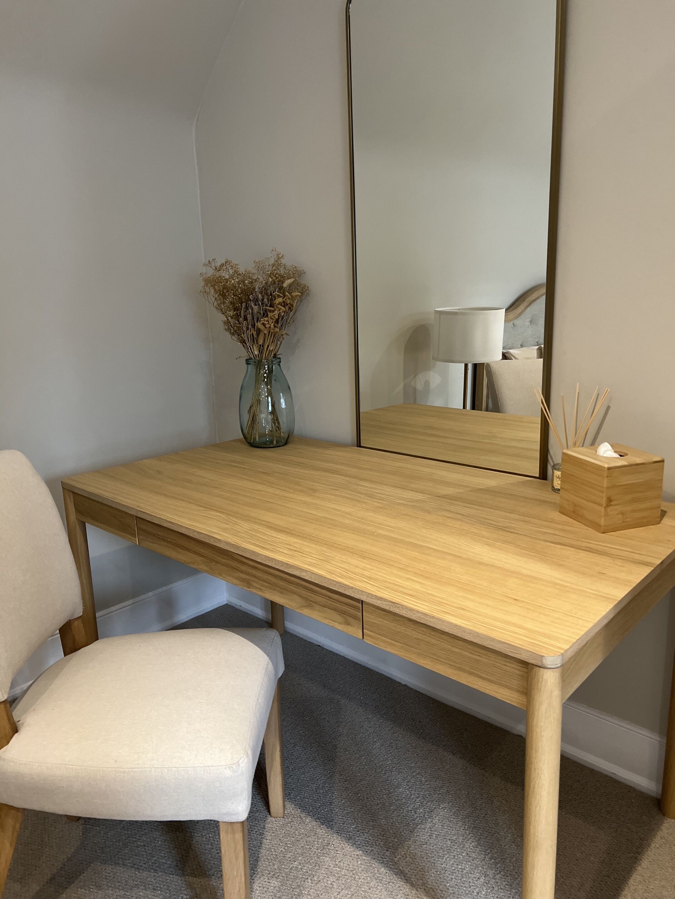 A wooden desk with a mirror, a vase with dried flowers, a tissue box, and a chair.