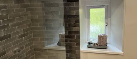 A bathroom with a glass shower enclosure, a bathtub, a window with a view, and a shelf with toiletries.