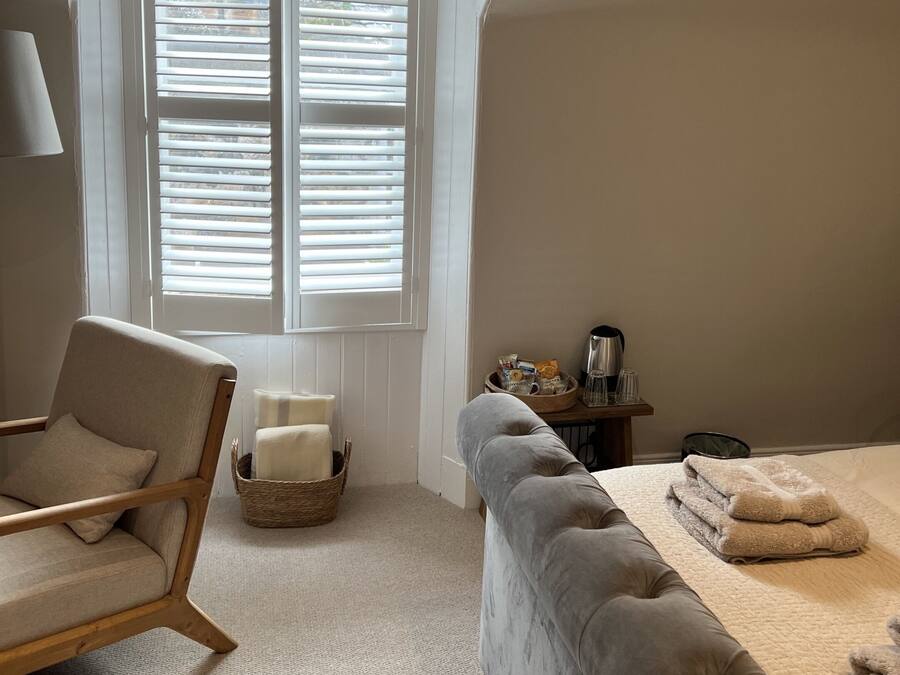 A bedroom with a large window, a bed with folded towels, a chair, a bedside table with a lamp, and a basket.