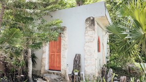 Edifici rústic amb una porta vermella, parets de pedra i un jardí ple de plantes i arbres diverses.