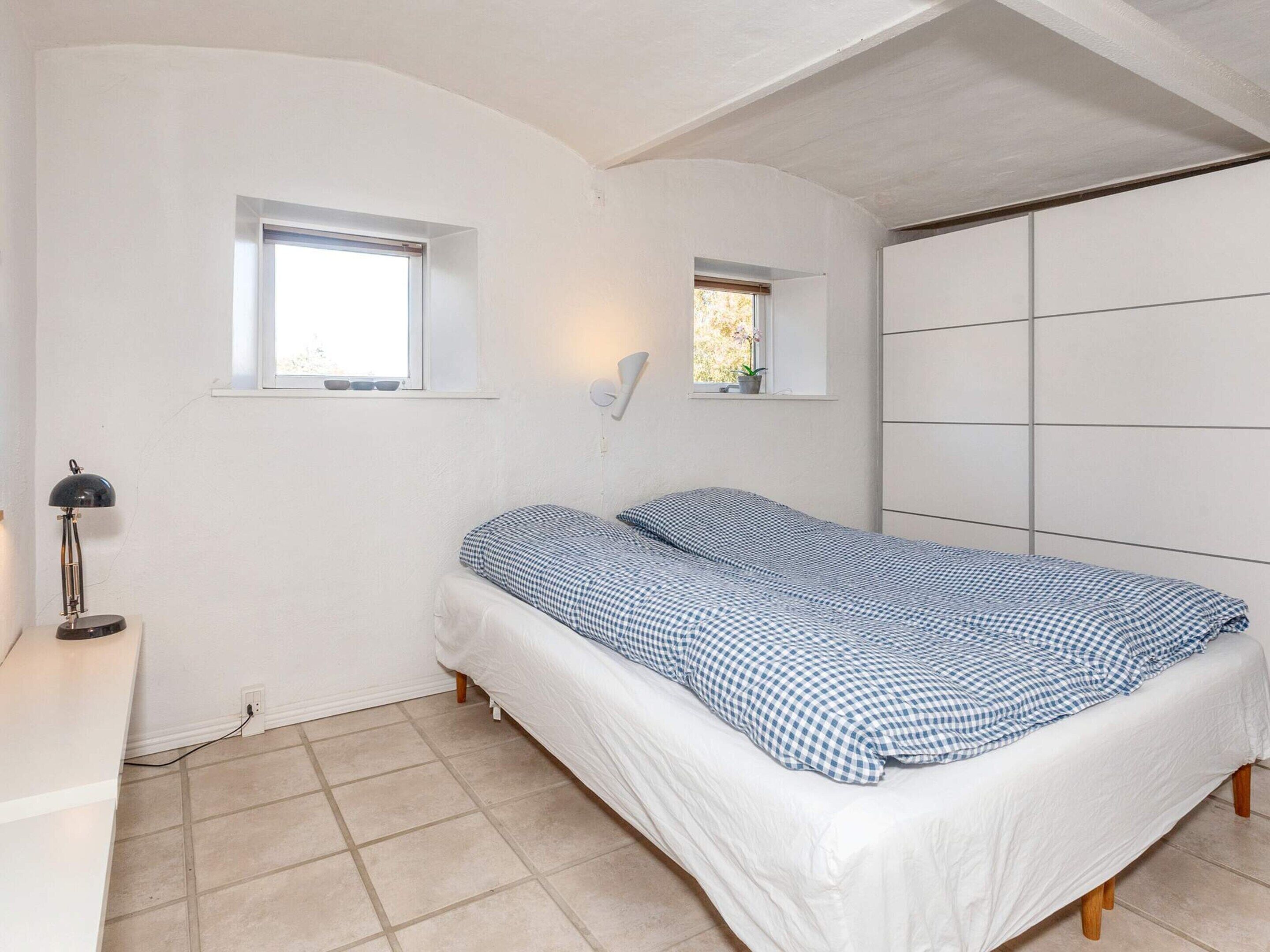 Un dormitorio con cama, dos ventanas, un escritorio con lámpara y un armario.
