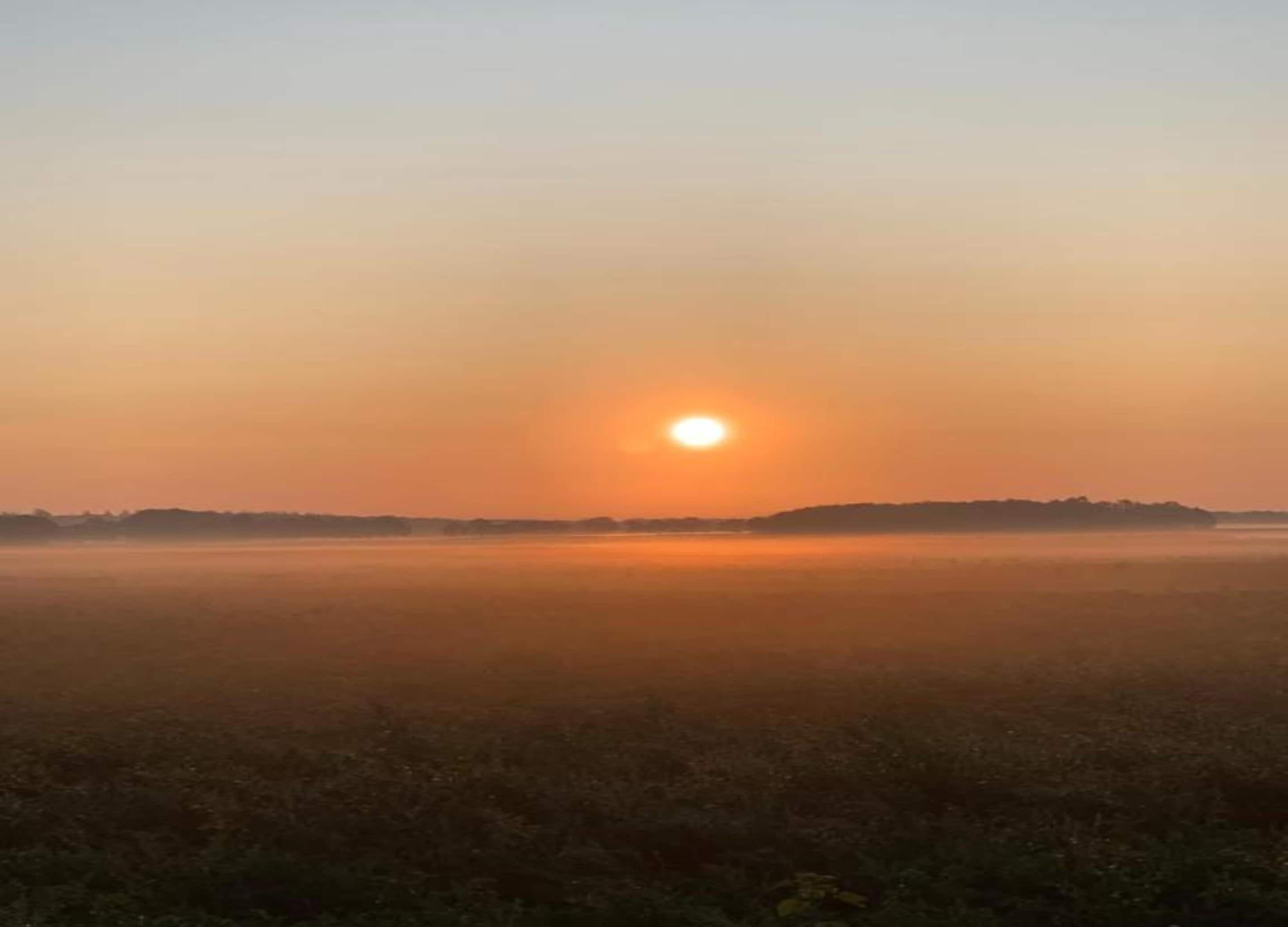 Een zonsopgang boven een vlak landschap met een nevelige horizon.