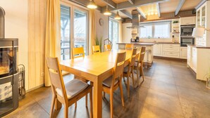 Cozinha moderna com vigas de madeira, área de jantar com mesa e cadeiras grandes e vista para o exterior por grandes janelas.