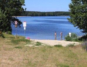 A lakeside view with a sandy beach, a metal railing, and two people standing by the water. - Ferienhaus am Oase am Vilzsee (Mirow)