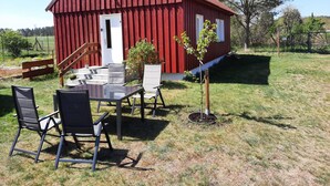 A red barn with a white door, a small window, and a staircase leading up. There is a table with chairs outside, and a young tree planted in the grass. - Ferienhaus am Oase am Vilzsee (Mirow)