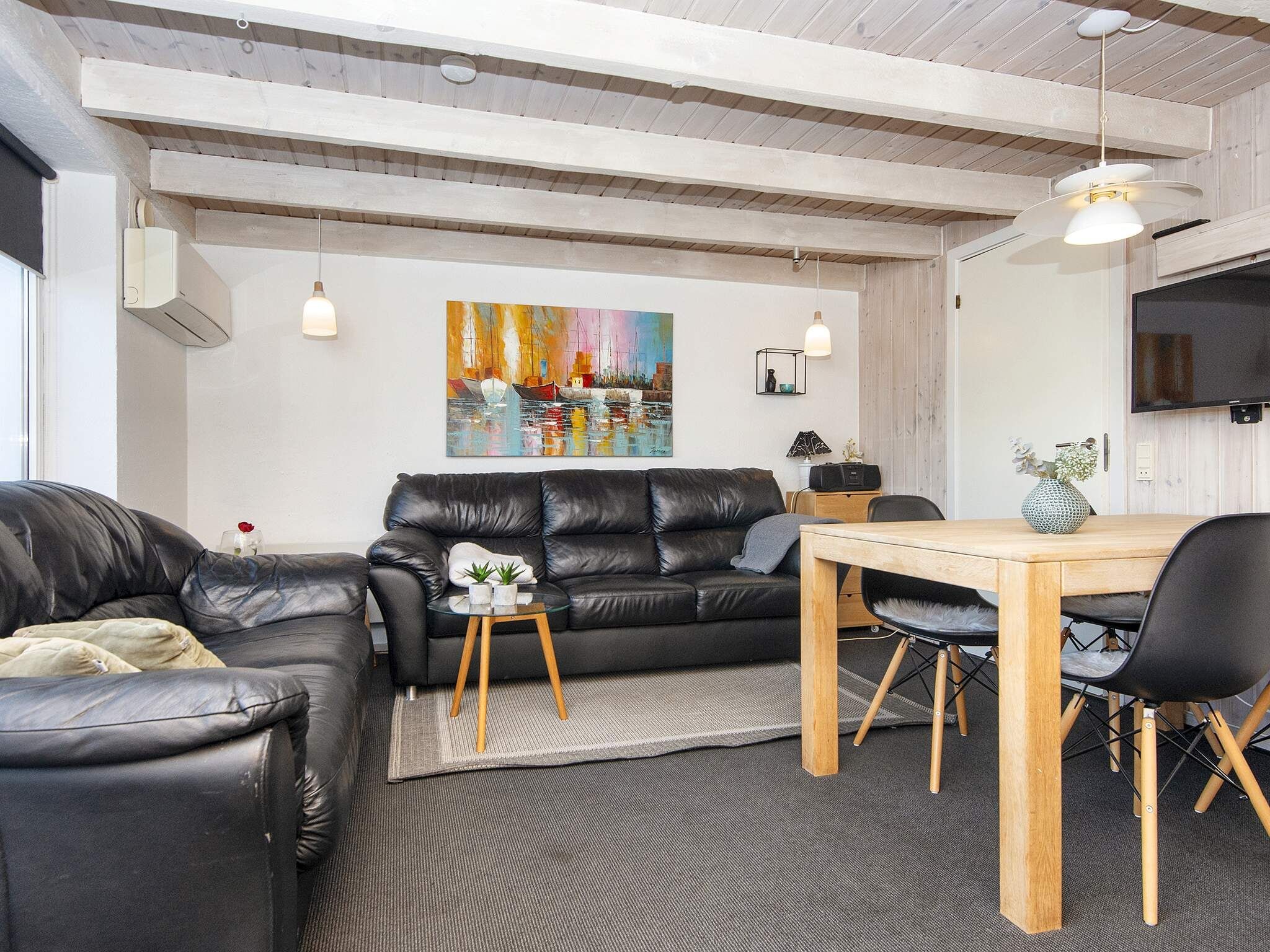 Una sala moderna con un sofá de cuero negro, una mesa de comedor de madera con sillas, un cuadro en la pared y un televisor de pantalla plana sobre la mesa.
