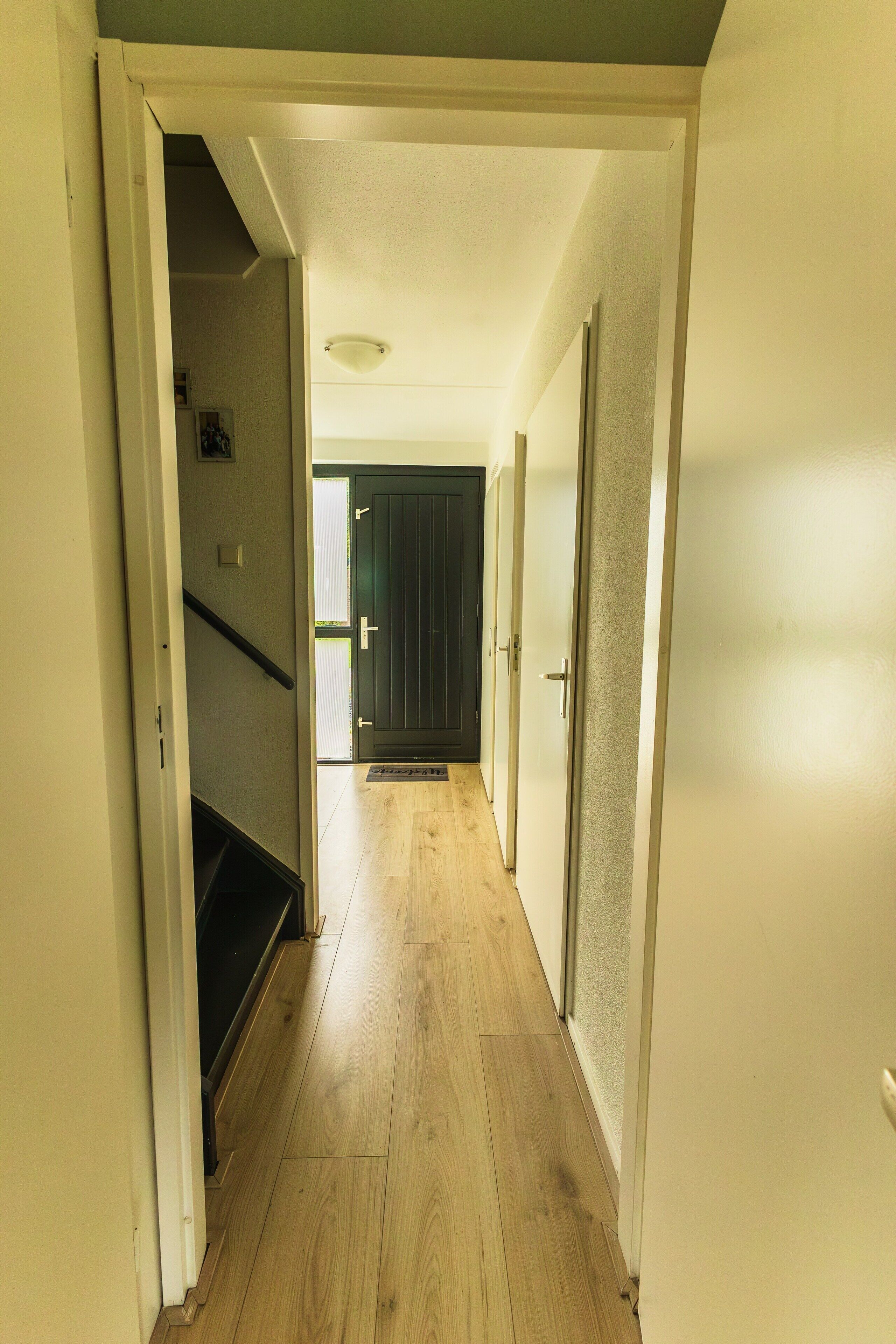 A hallway with wooden flooring, a staircase, and a door leading to another room.