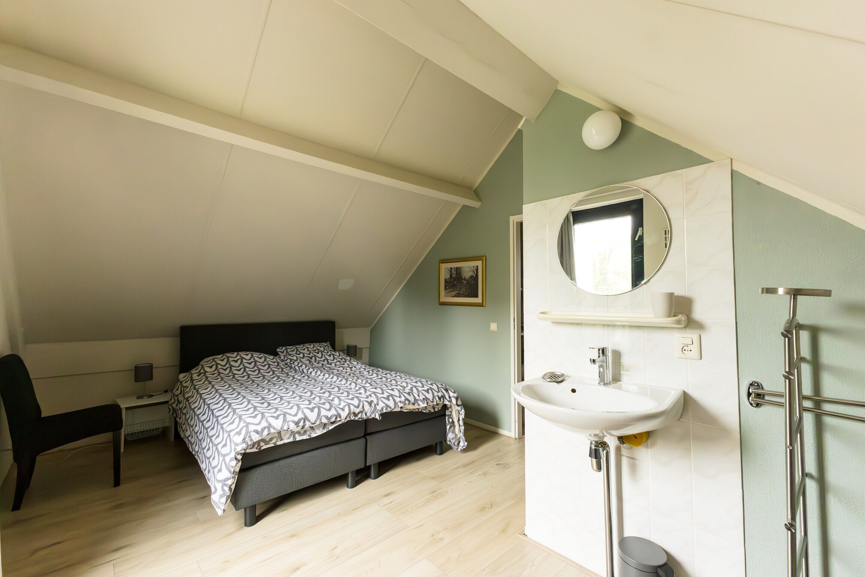 A bedroom with a bed, a desk, a chair, a bedside table, a mirror, a sink, and a shower.