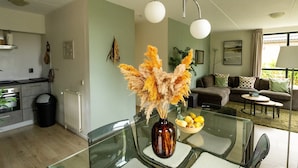 A modern kitchen with a dining area, a living room with a sofa and a coffee table, and a vase with dried flowers on the dining table.