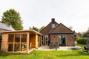 Et murstenshus med terrasse, en trækonstruktion med glasdøre og en græsplæne.