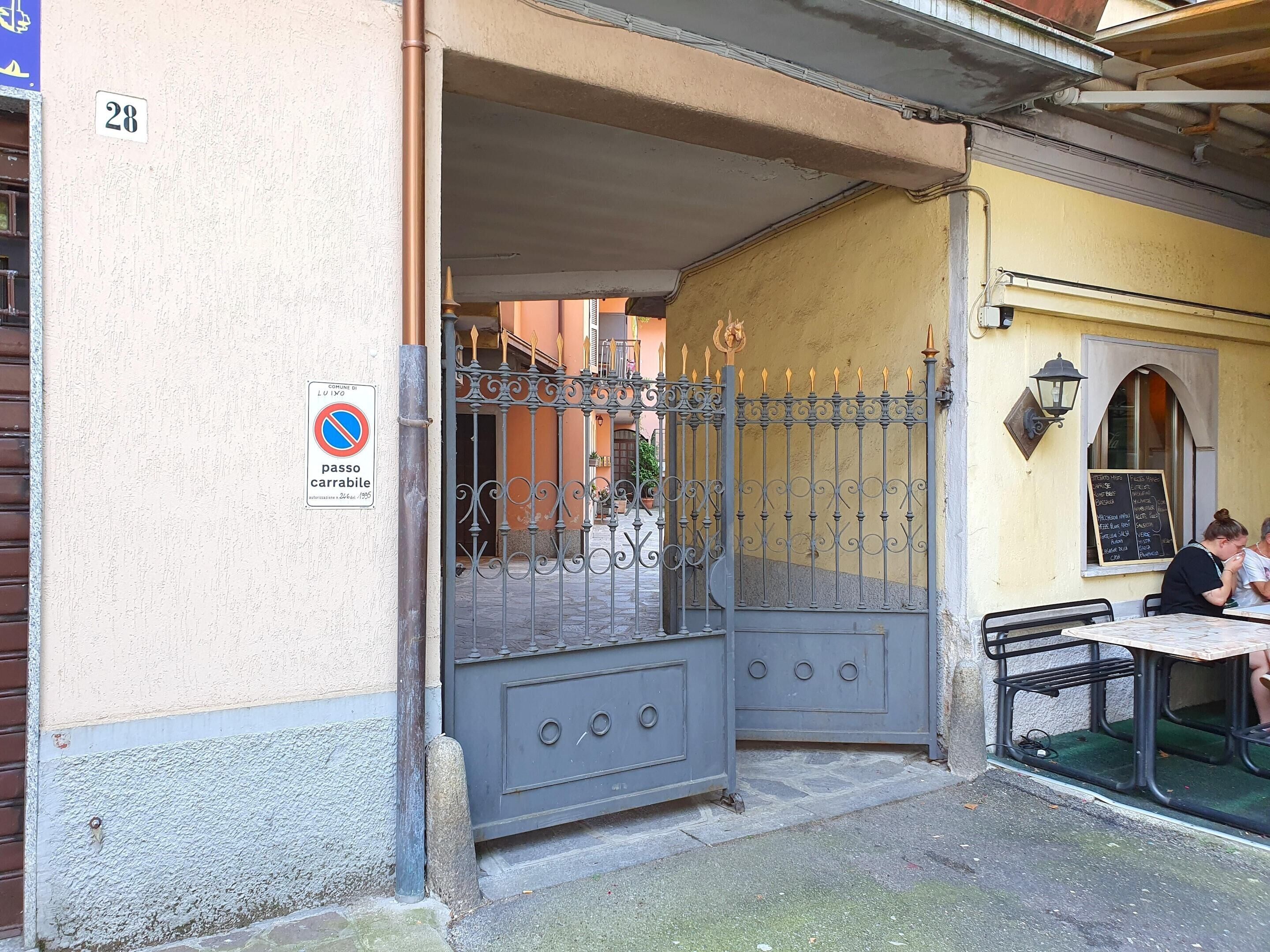 Une entrée fermée par un portail mène à un immeuble sur lequel est apposé un panneau indiquant qu’il est interdit de se garer. On y trouve un banc à l’extérieur et une table où sont assis deux personnes.