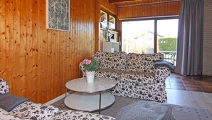 Een knus woongedeelte met houten wanden, een bloemenbank, een ronde salontafel met een bloevas en uitzicht op het landschap door een groot raam.