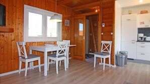 Een houten interieur met een eethoek, witte stoelen, een tafel, een keuken met witte kastjes en een plafondlamp.