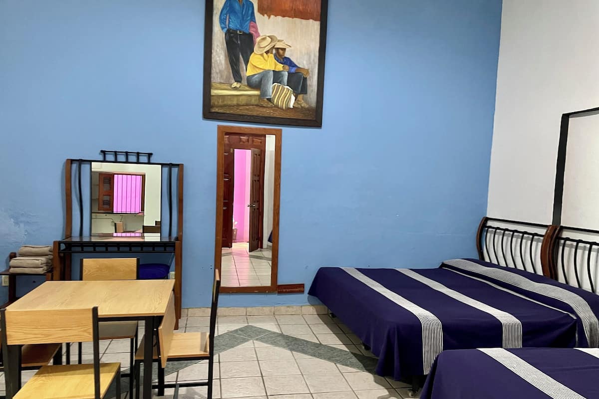 A room with a desk, two single beds, a chair, a mirror, and a painting on the wall.