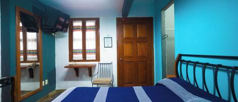Un dormitorio con cama, puerta de madera, ventana con cortinas, una mesita y una silla.