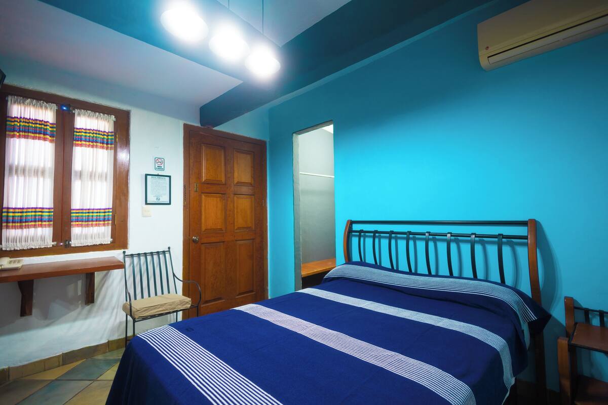 A bedroom with a bed, a wooden door, a window with curtains, and an air conditioning unit.