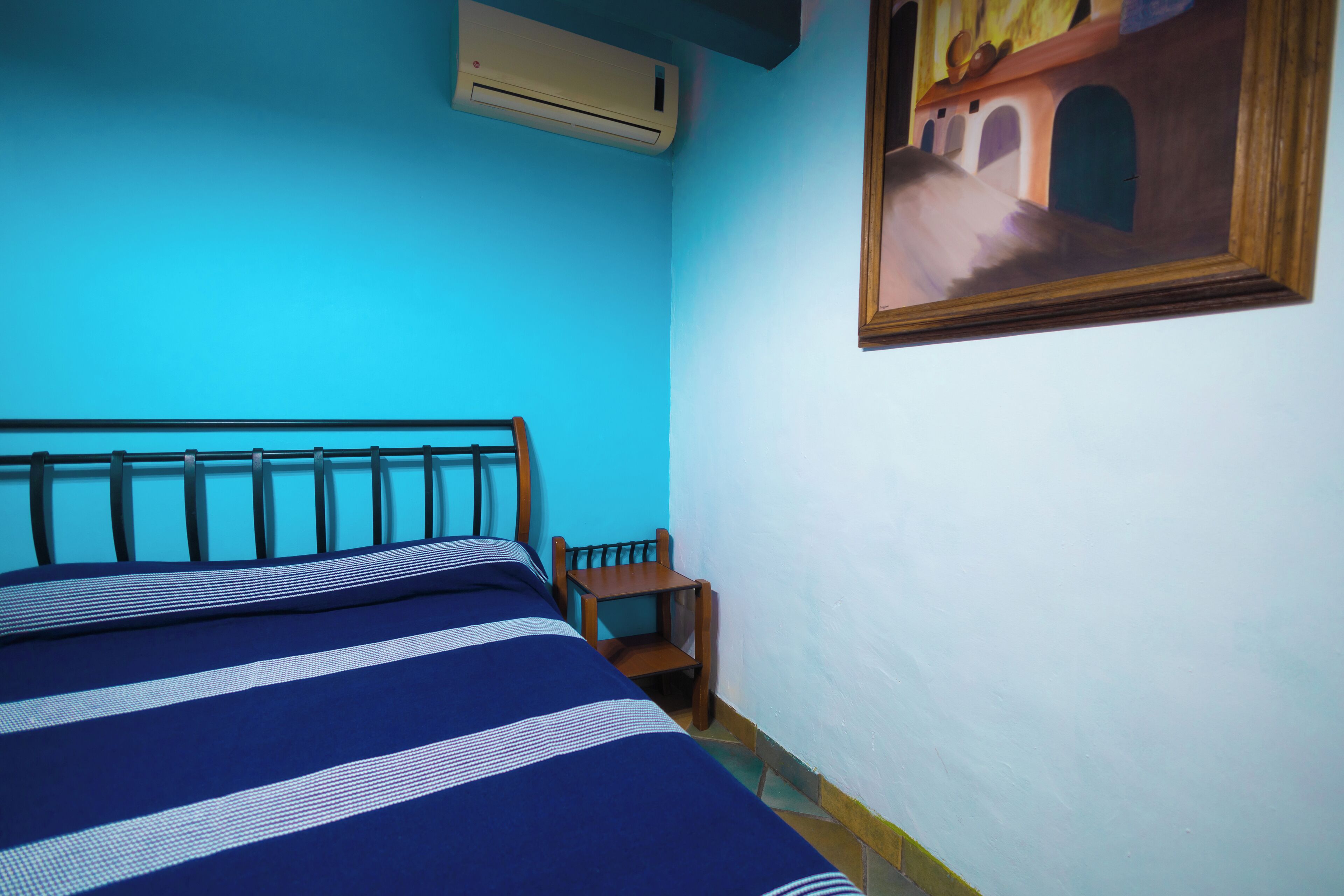 Ein Zimmer mit einem Bett, einem kleinen Holzstuhl, einer Klimaanlage und einem Bild an der Wand.