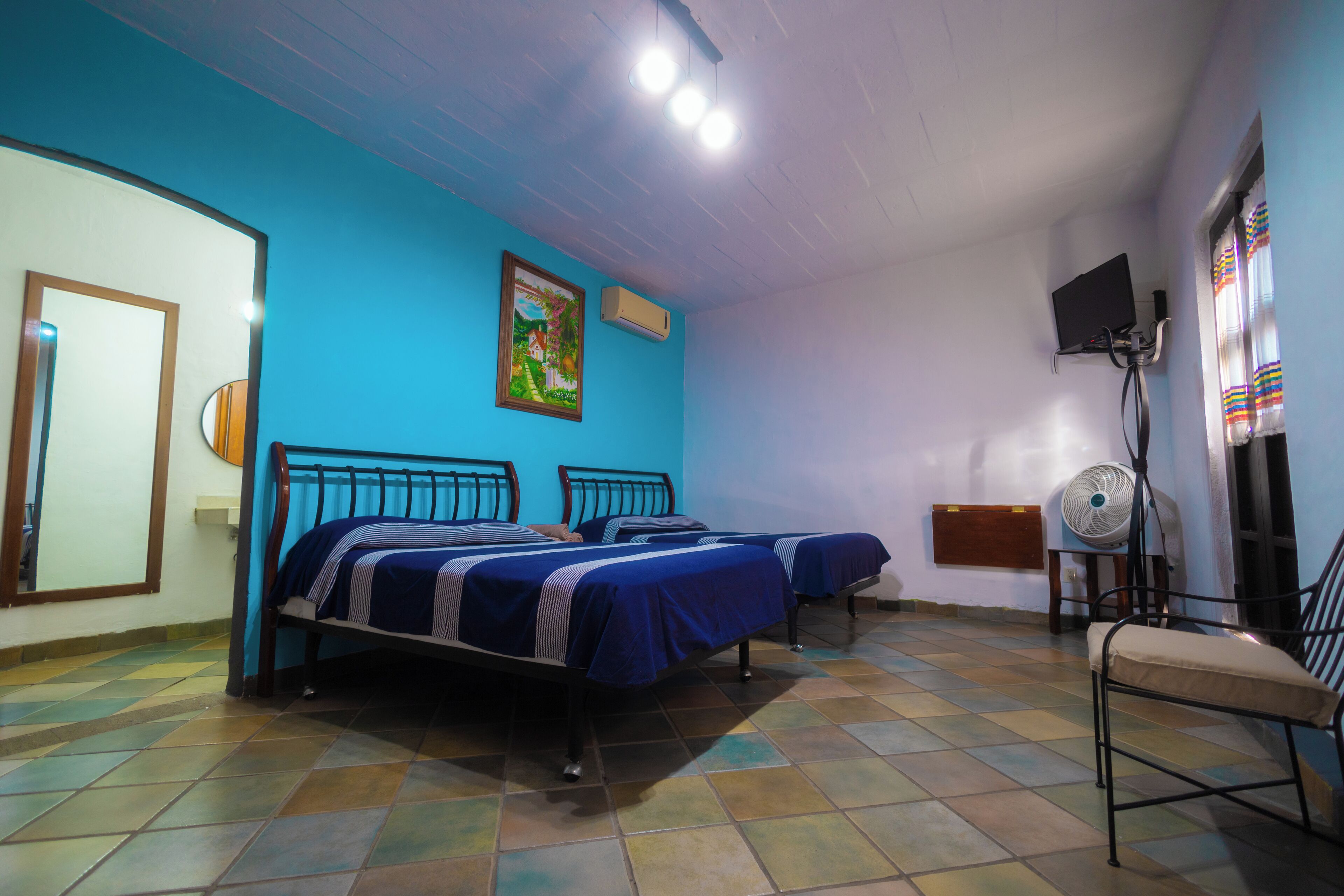 A room with two beds, a chair, a fan, and a painting on the wall.