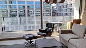 Un salon moderne avec un canapé, un fauteuil en cuir noir, une petite table et un balcon offrant une vue sur un immeuble de grande hauteur.