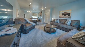 A spacious living room with a large sofa, a coffee table, and a TV. There is a kitchen in the background with a dining area. - -:- Cowboy Casa -:- Modern New Construction - 3BR+  (Cody)