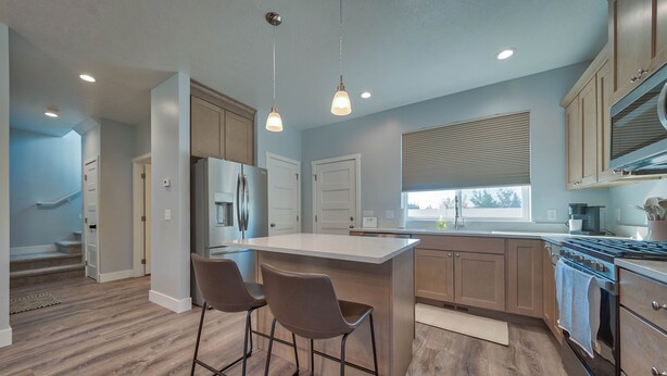 A modern kitchen with a central island, bar stools, wooden cabinets, stainless steel appliances, and a window with blinds. - -:- Cowboy Casa -:- Modern New Construction - 3BR+  (Cody)