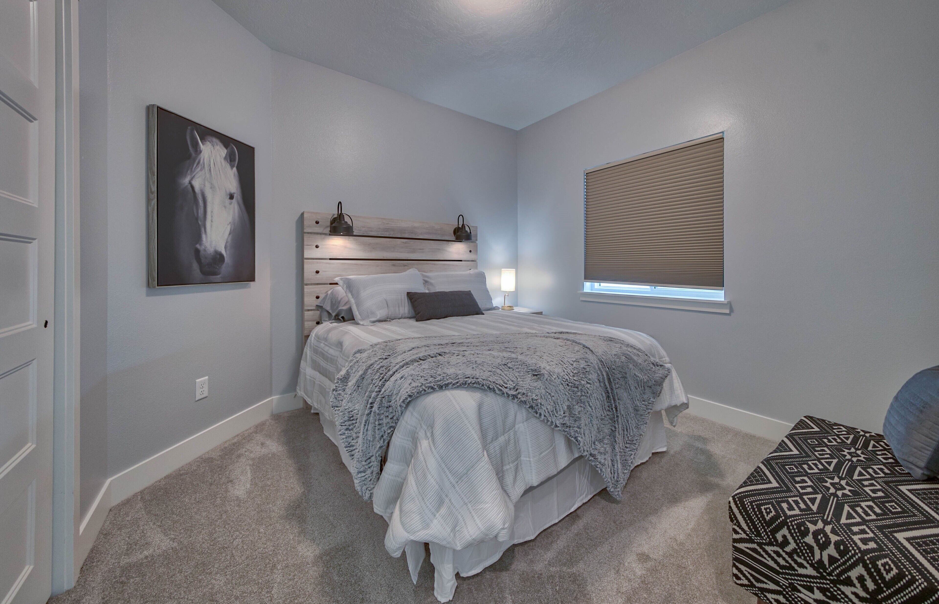 Une chambre à coucher comprenant un lit, une tête de lit, une fenêtre avec des stores, un luminaire fixé au mur et un tableau encadré.