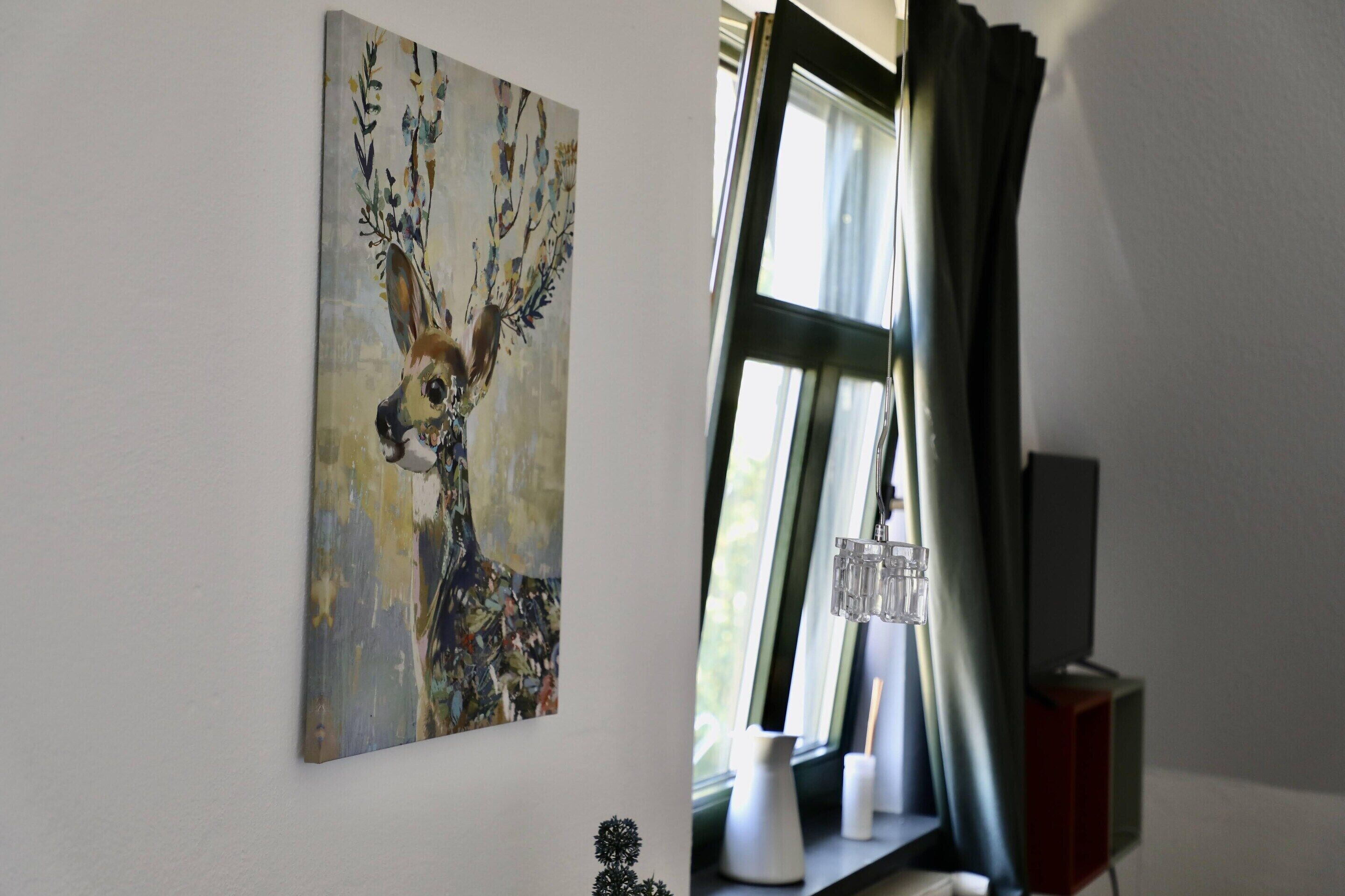 Une pièce avec un tableau représentant un cerf accroché au mur, une fenêtre avec des rideaux et une petite table sur laquelle se trouvent un vase et une bougie.