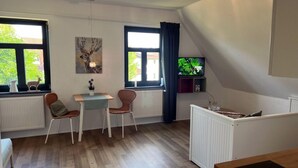 A room with a wooden floor, a dining table with chairs, a small white table, a radiator, a TV on a shelf, and a window with a curtain. - Studio in Dargun With Pool Near Lake Kummerow (Dargun)