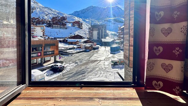 Blick aus einem Fenster mit Holzboden auf eine verschneite Bergstadt mit Gebäuden und blauem Himmel.