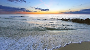 Una playa con olas que acarician suavemente la orilla, un cielo despejado y una puesta de sol en la distancia.