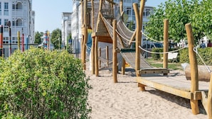 Una estructura de juego de madera con un puente de cuerda y un tobogán, rodeada de arena y vegetación, con edificios al fondo.