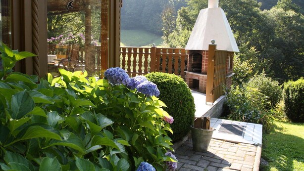 Eine Terrasse mit einem Backofen aus Ziegelsteinen, umgeben von üppiger Vegetation und Blick auf einen See.
